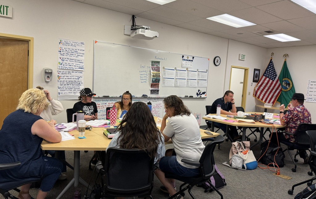 A group of people sit around tables in a classroom. Papers, books, and flags are visible.