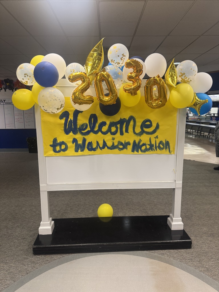 A white board on a stand with a yellow banner saying "Welcome to Warrior Nation." Decorated with balloons and foil letters "2030."