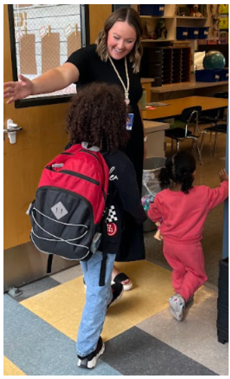 A woman in black stands in a classroom, guiding a young child with a pink outfit and another with a red backpack.