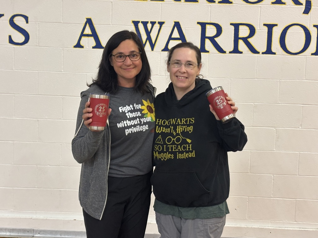 Two women stand next to each other, holding cups. One wears glasses and a gray hoodie, the other a black hoodie. The wall behind them reads "S A WARRIOR".