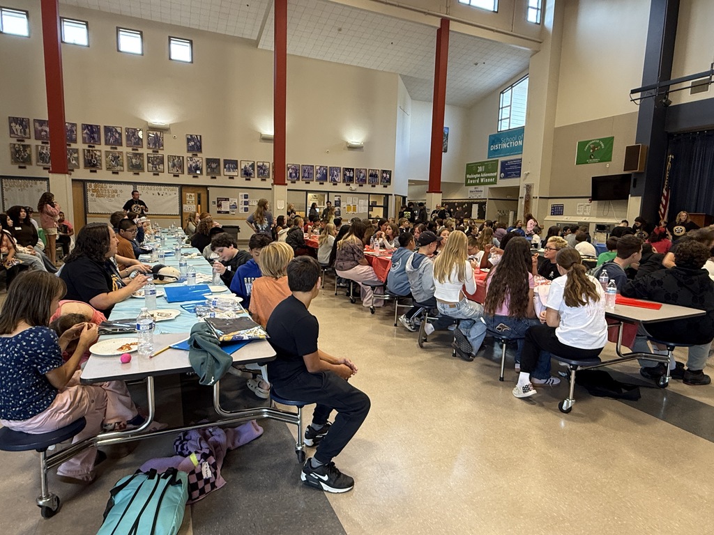 Indoor cafeteria with tables, chairs, and people sitting and standing. Several tables have food items on them.