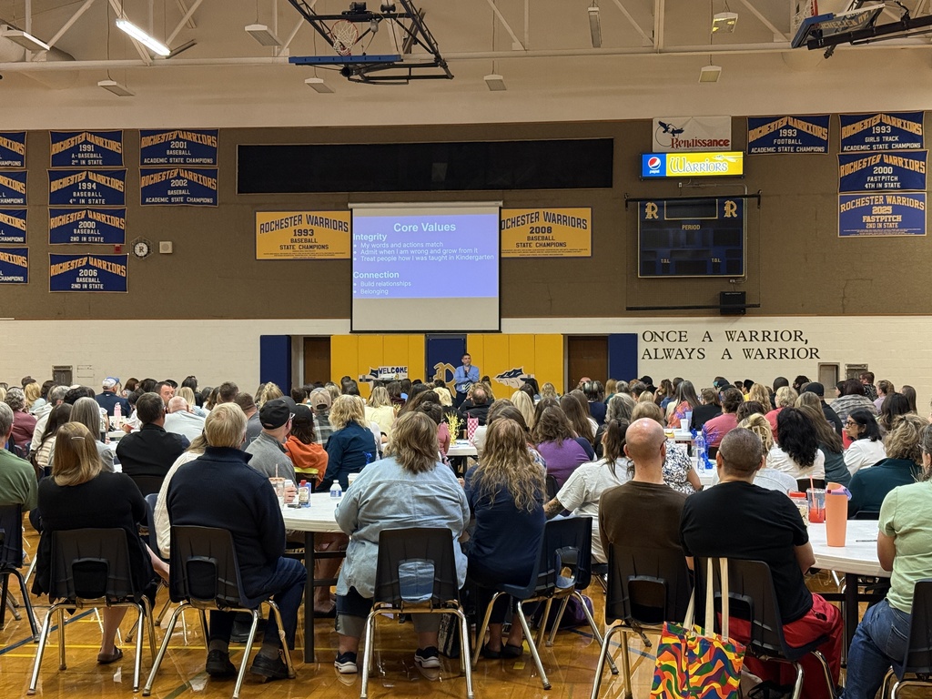 A large room with tables and chairs, many people sitting. A projector screen displays content.