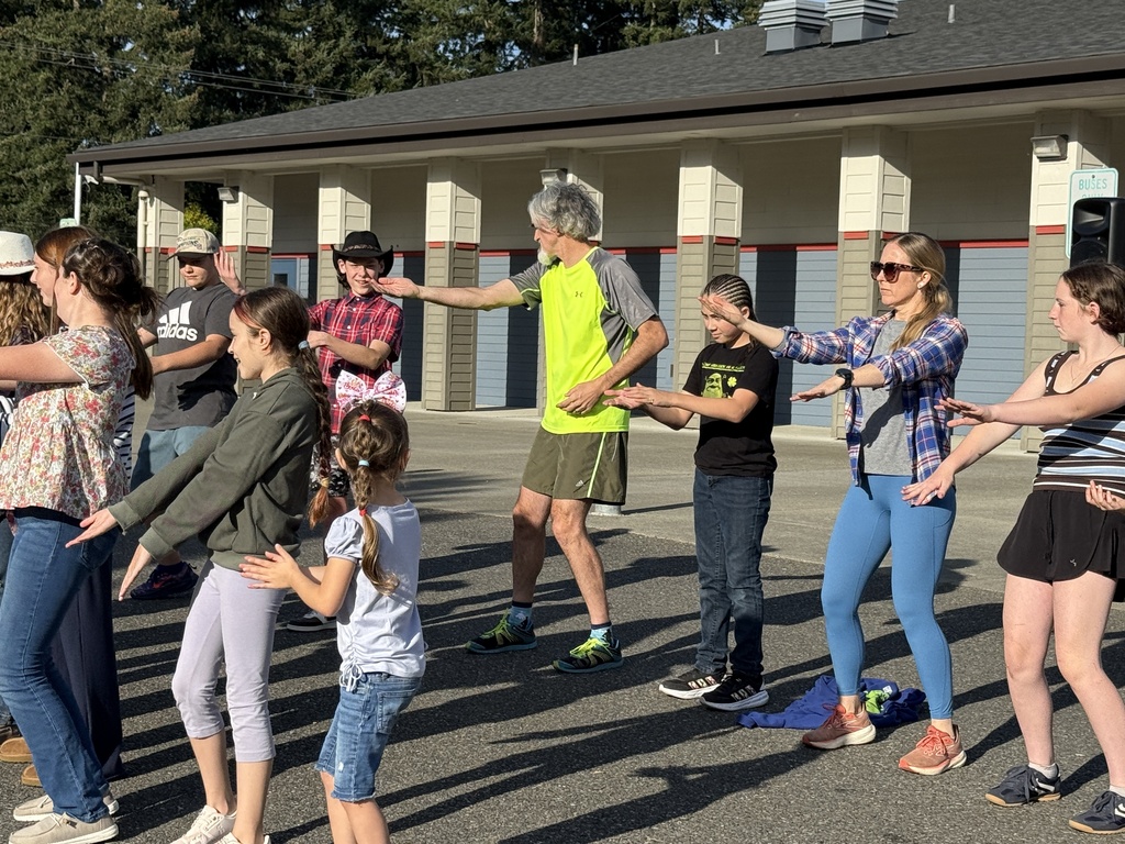 Group of people in a parking lot. Two adults in the center lead a line of children in a dance.