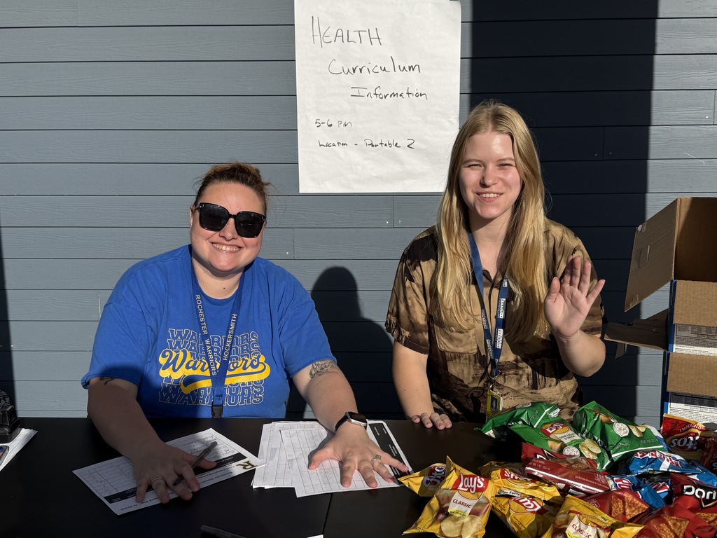 Two people sit at a table, smiling. A sign reads "Health Curriculum Information". Various snacks are on the table.