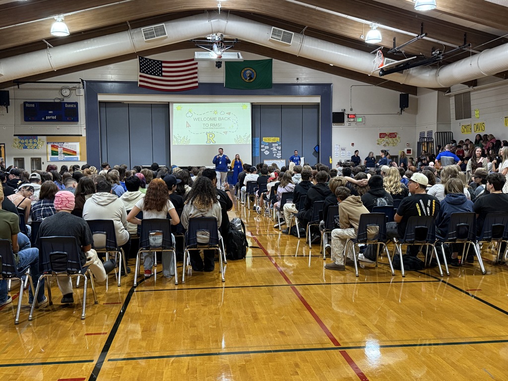 Gymnasium with a projector screen at the front; many people seated, some standing; two flags on the wall.