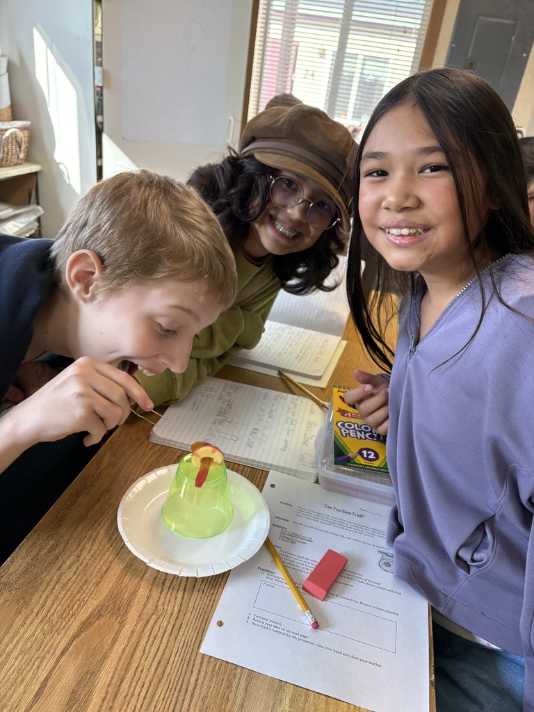 A boy and a girl are leaning over a table with a yellow object. Another child stands beside them. Papers, a pencil, and a rubber are on the table.
