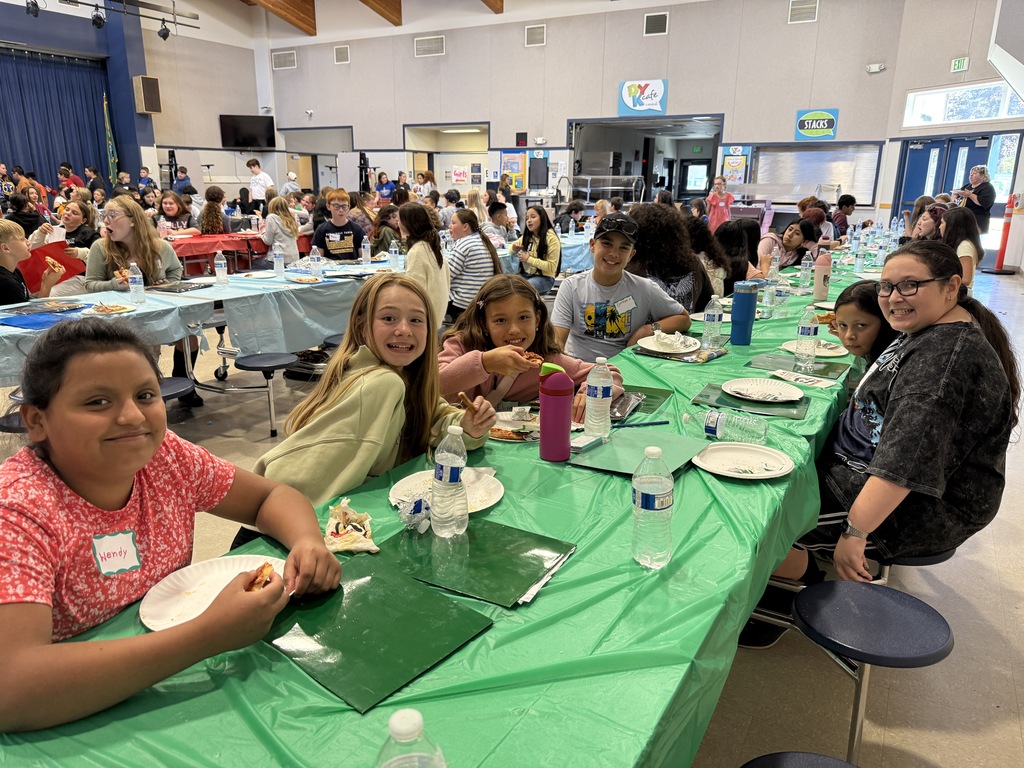 Several children sit at a long table with plates, water bottles, and napkins in a school cafeteria.