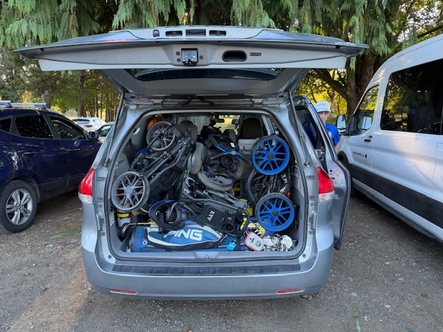 Car with open trunk filled with bikes, helmets, and sports equipment. Trees and other vehicles visible in the background.