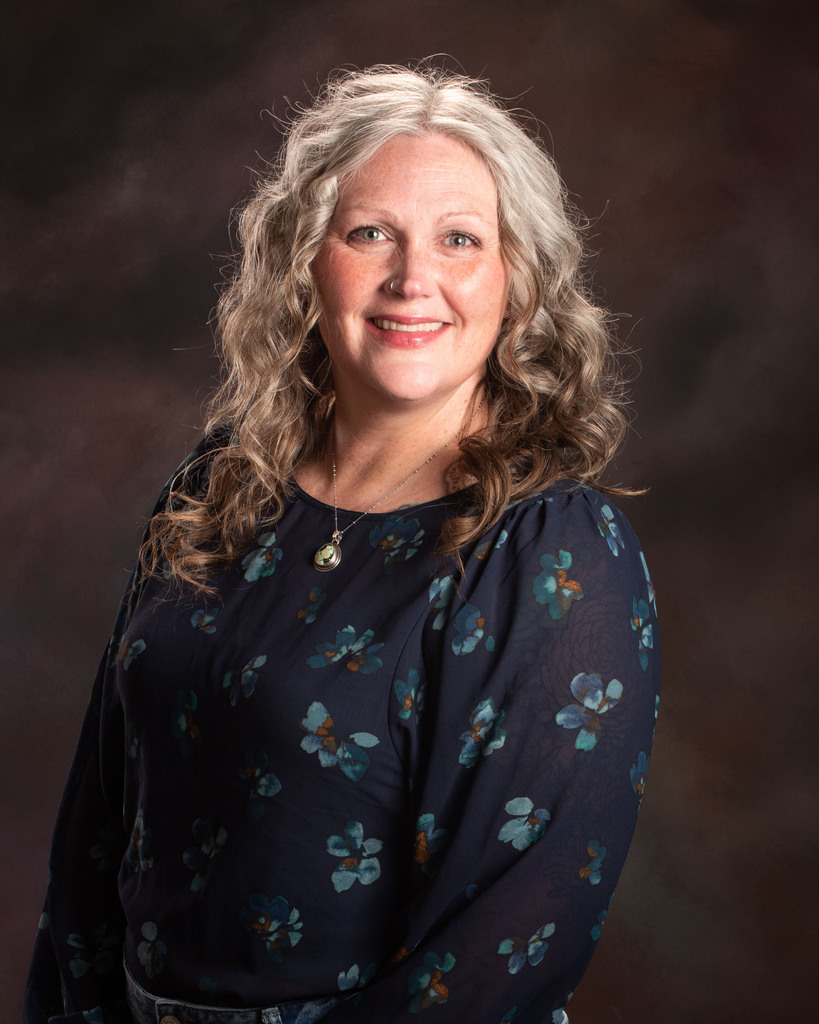 A woman with white curly hair wearing a floral shirt stands against a dark brown background.