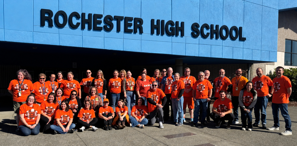 A group of people wearing orange shirts poses in front of the Rochester High School building.