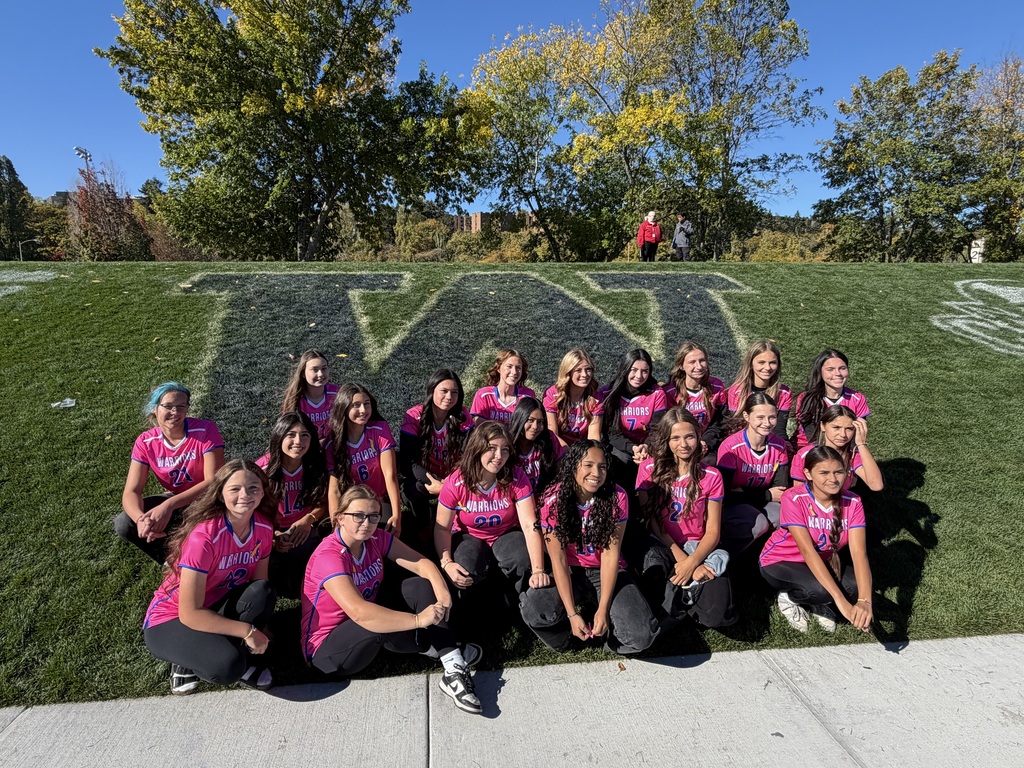 A team in pink shirts poses on a grassy field, with a large "W" and trees in the background.