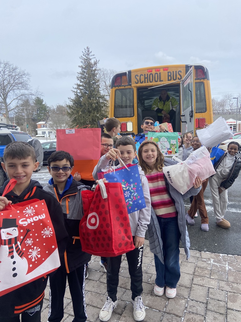 Female and Male 4th grade students with presents on hand  while posing in front of the school bus that will take them to drop off their giving tree donations 