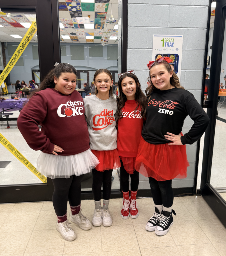 four students dressing with coca cola them eoutfits- A Refreshing Take on Halloween! These 6th graders brought some creativity to our Halloween Social with their Coca-Cola-inspired costumes, featuring Classic Coca-Cola, Diet Coke, Coke Zero, and Cherry Coke!