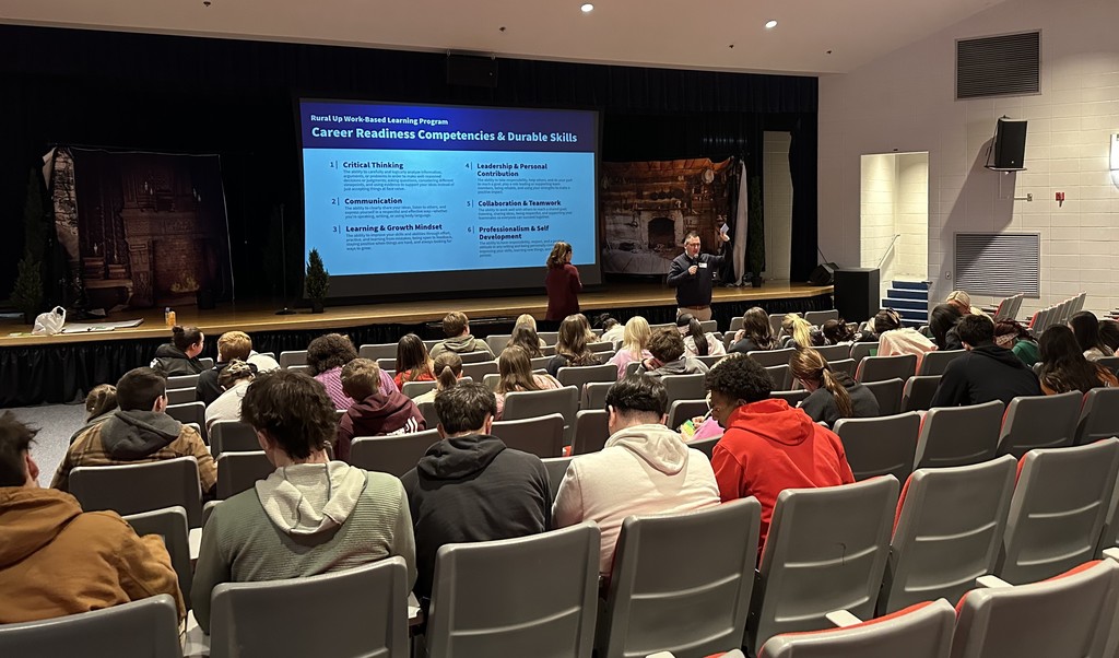 High school students sit in a school auditorium.   A speaker stands in front of the room with a presentation visible. 