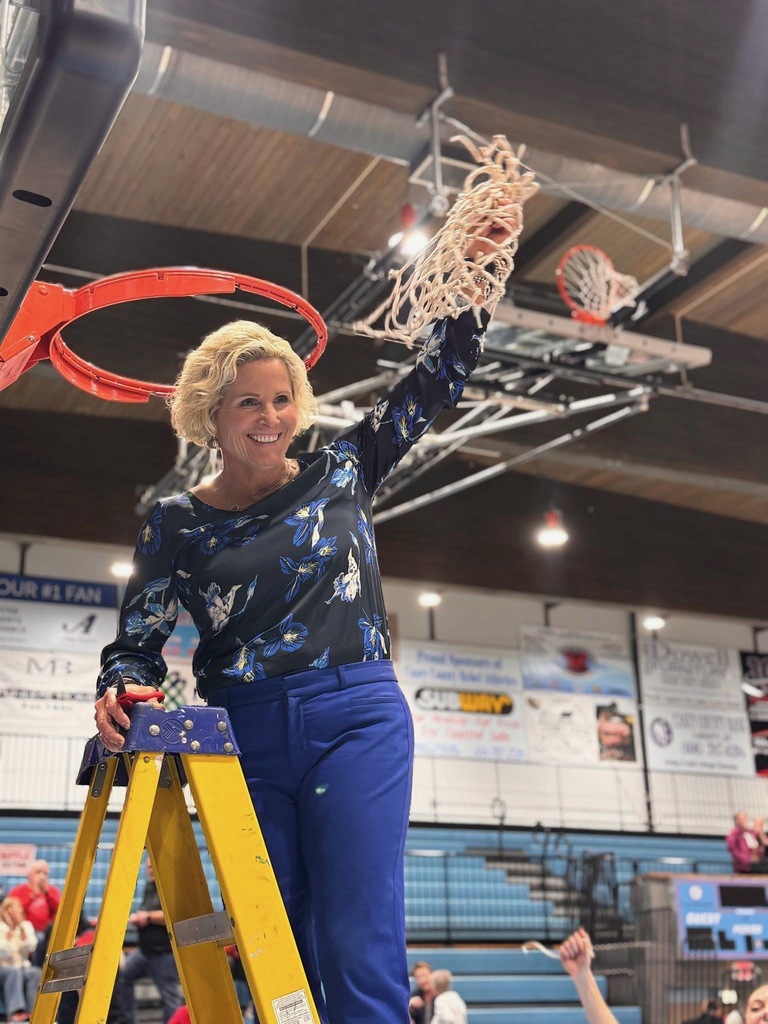 Coach Noble stands on a yellow ladder holding a basketball net over her head. 