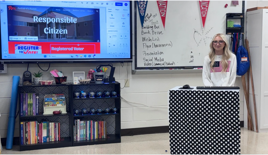 A high school aged female stands behind a podium covered in black and white polka dots.  She  has long blonde hair and is wearing a white sweater with an American flag on the front.  