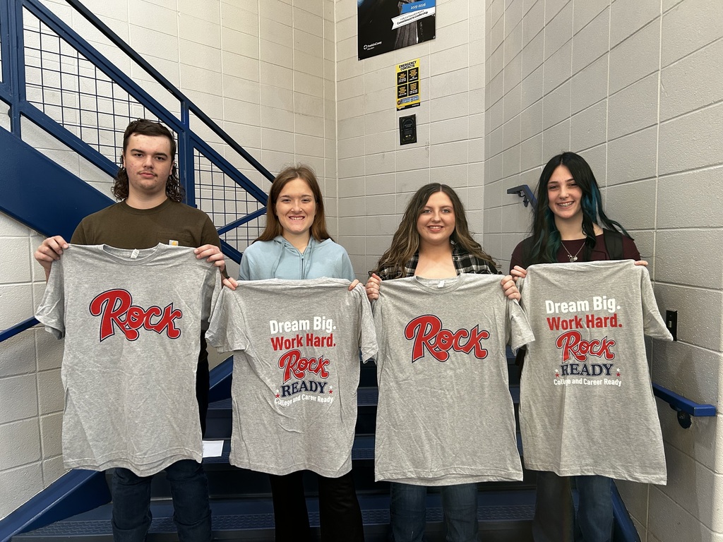 Four students stand at the foot of the stairs holding up t-shirts.   