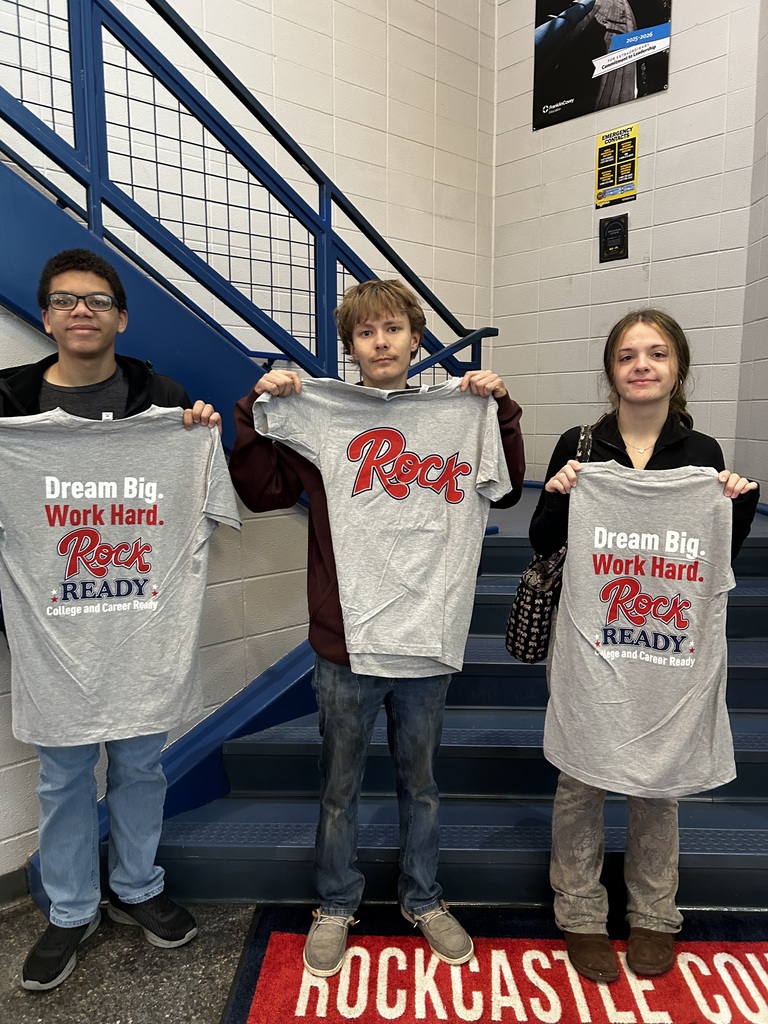 Three high school students, two males and a female , stand at the foot of steps holding up t-shirts. 