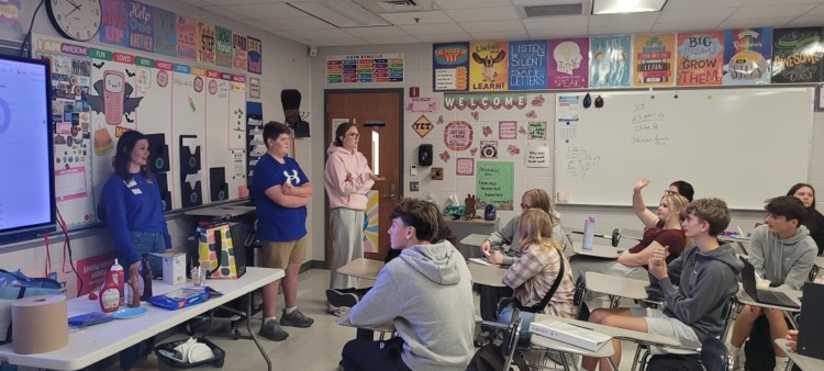 Students sit with back to the camera as other students stand in front of the room. A math term appears on the large screen  
