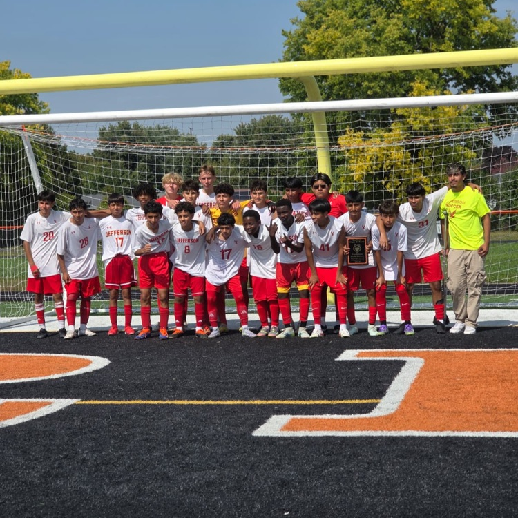 Jefferson High School freshman boys soccer team poses together on the field holding their championship trophy after winning the Harlem Tournament. The players are in red uniforms with gold accents, smiling and celebrating as a group.