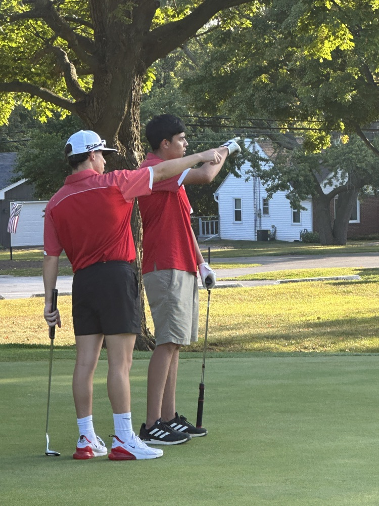 Jefferson golfers in red polos and khakis competing on the course — teammates discussing a putt on the green and a player taking a swing off the tee box.