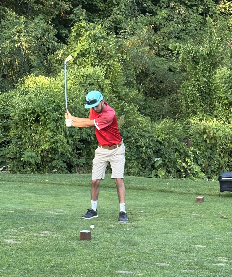 Jefferson golfers in red polos and khakis competing on the course — teammates discussing a putt on the green and a player taking a swing off the tee box.