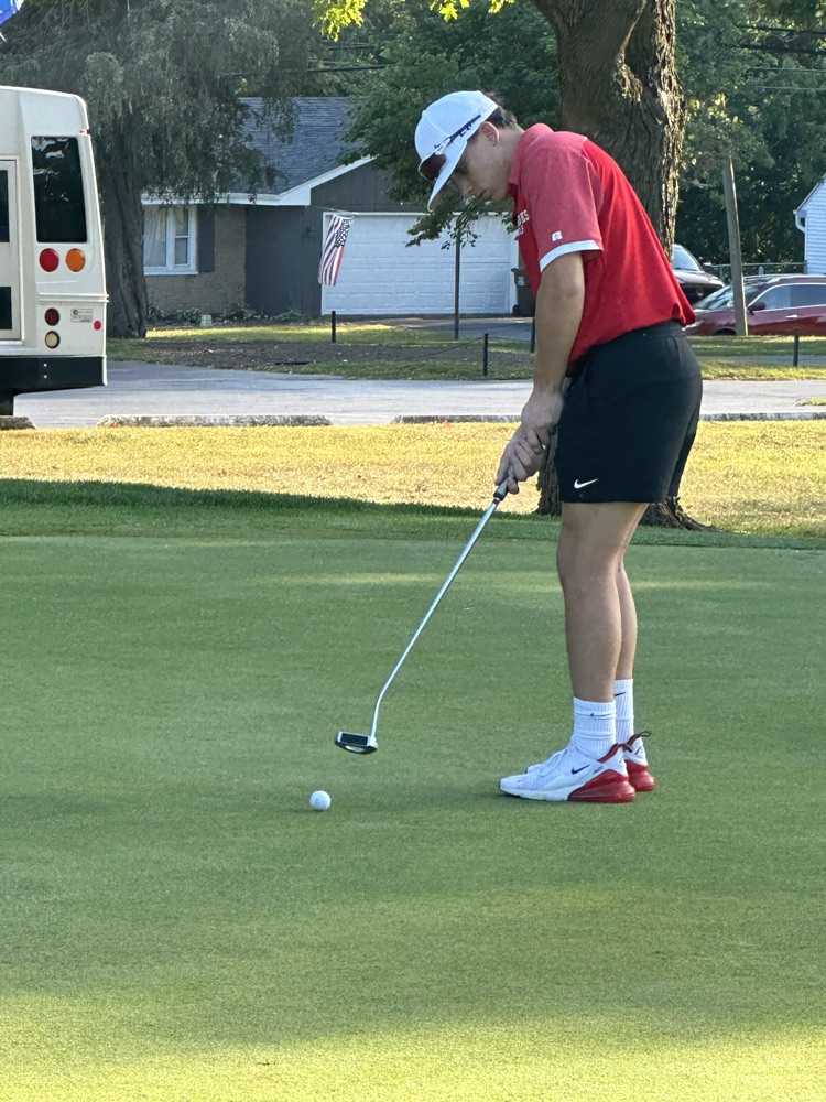 Jefferson golfers in red polos and khakis competing on the course — teammates discussing a putt on the green and a player taking a swing off the tee box.