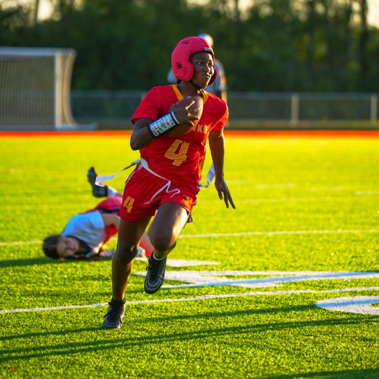 Alt text: A Jefferson High School football player in a red uniform and helmet runs with the ball down the field as a defender dives and falls behind him. The sun sets in the background, highlighting the green turf.