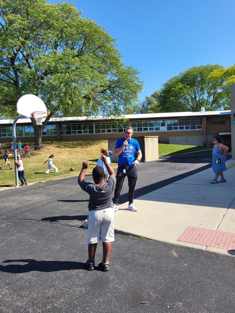 Man speaking on microphone and students playing on blacktop.