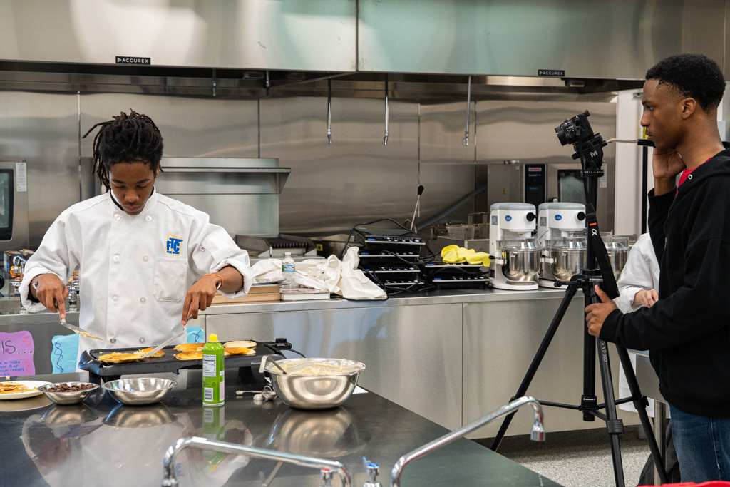 Student is cooking pancakes and another student is filming them.