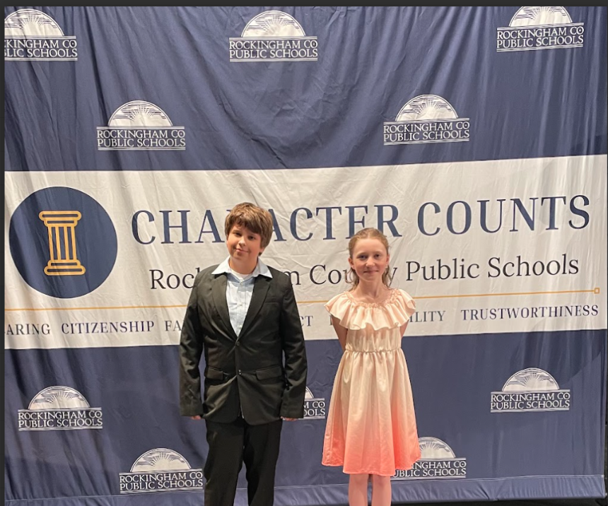 A blue banner in the background with a yellow pillar and the logo to Rockingham County Public Schools is randomly placed around the blue background. Across the middle of the blue background are words that say Character Counts Rockingham County Public Schools Caring Citizenship respect fairness responsibility trustworthiness. Standing in front of the blue background is a boy wearing a black jacket and black pants with a blue shirt. He has brown hair and is smiling. The girl is wearing a light pink ombre style dress and her blonde hair is pulled back from her face. She is smiling