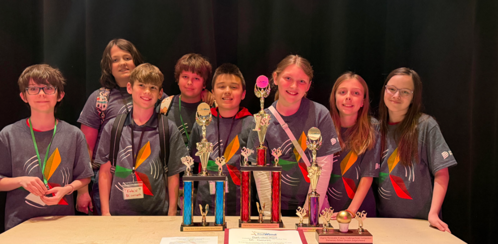 7 fifth grade students. 4 boys on the left and 3 girls on the right. All children are wearing gray shirts with a yellow, red and green windmill on it. The students are standing in front of a brown table with 3 big trophies and one small trophy that they have won. All students are smiling. One trophy is gold and blue, one is red and gold and one is purple and gold and one is just gold.
