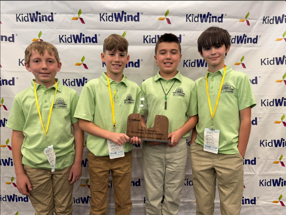 4 boys in green shirts and khaki pants with yellow lanyards holding a wooden trophy while standing in front of a backdrop that is white and has the word kidwind and several green, yellow and purple pinwheels