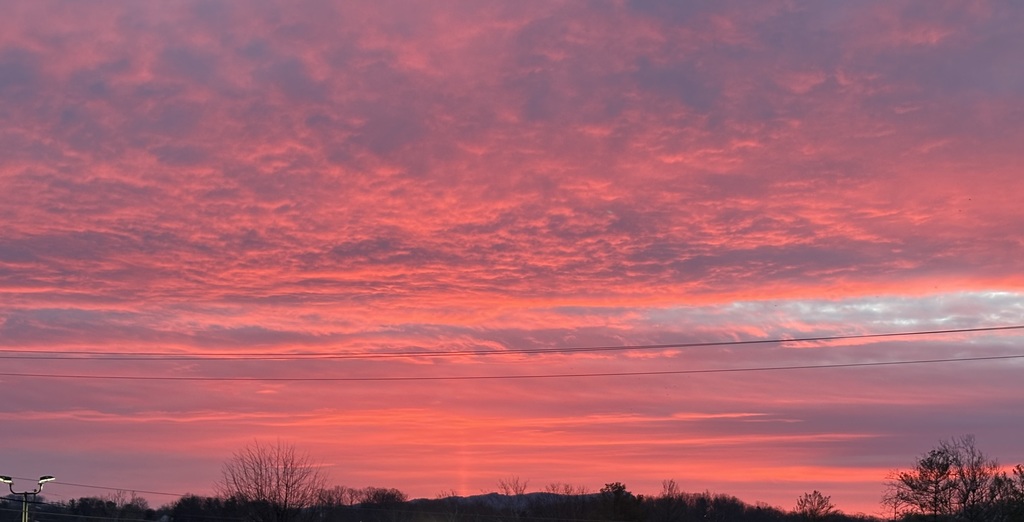 A vivid sunset fills the sky with shades of pink, red, and orange, layered with soft, textured clouds. Dark silhouettes of trees line the horizon, with low hills in the distance. Power lines stretch horizontally across the scene, contrasting against the colorful sky.