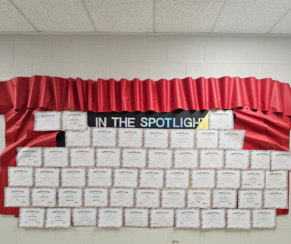 A photo of a school bulletin board titled "IN THE SPOTLIGHT" in shimmering letters. The board is framed by red paper curtains and is completely covered with rows of over 40 individual "Certificate of Completion" papers, showcasing the academic achievements of various students.