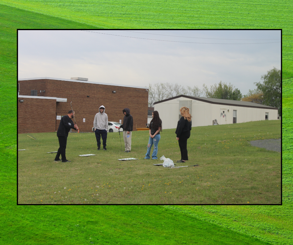 Five individuals are standing on a grassy field with buildings in the background. They appear to be practicing a sport.