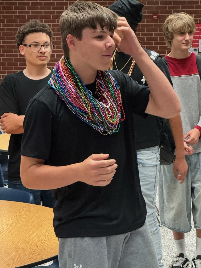 A few young men in a room with brick walls. One is wearing a colorful necklace.