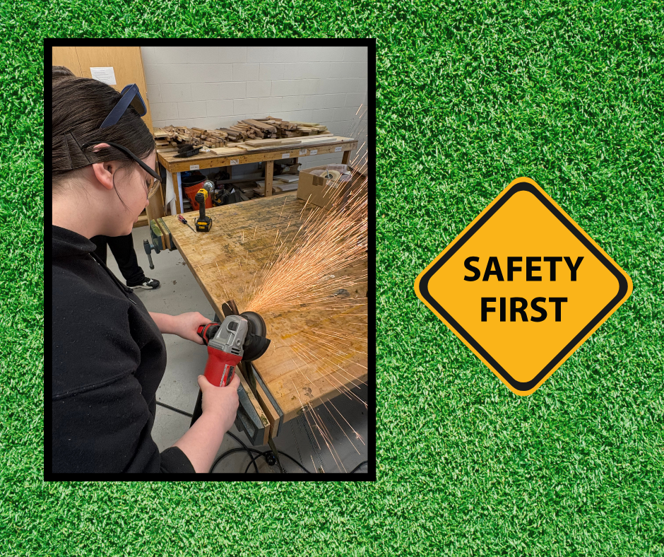 A woman in a workshop uses a grinder on a wooden table. A safety sign reading "Safety First" is placed in the foreground.