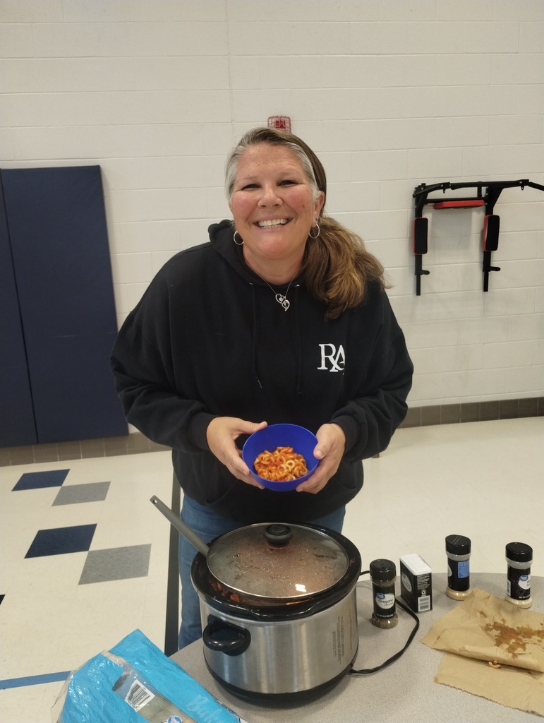 A woman in a black hoodie smiles, holding a bowl of food, with a crockpot and condiments on a table in a room.
