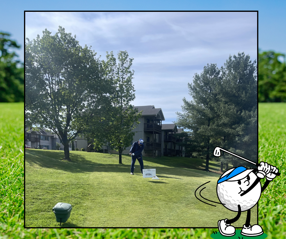 A golfer on a green, with a club in hand, surrounded by trees and a building. The golfer wears a hat and a blue shirt.