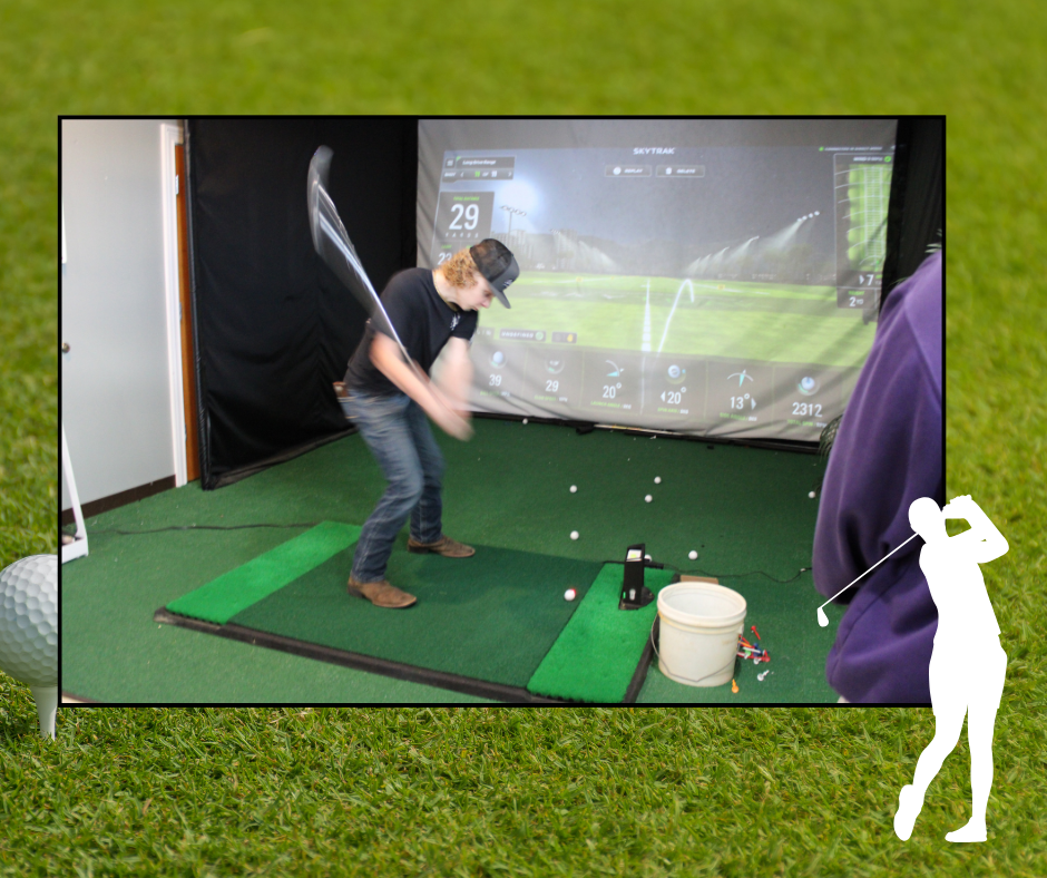 A golfer swings a club at a golf simulator with a large screen. Balls lie scattered on the green mat.
