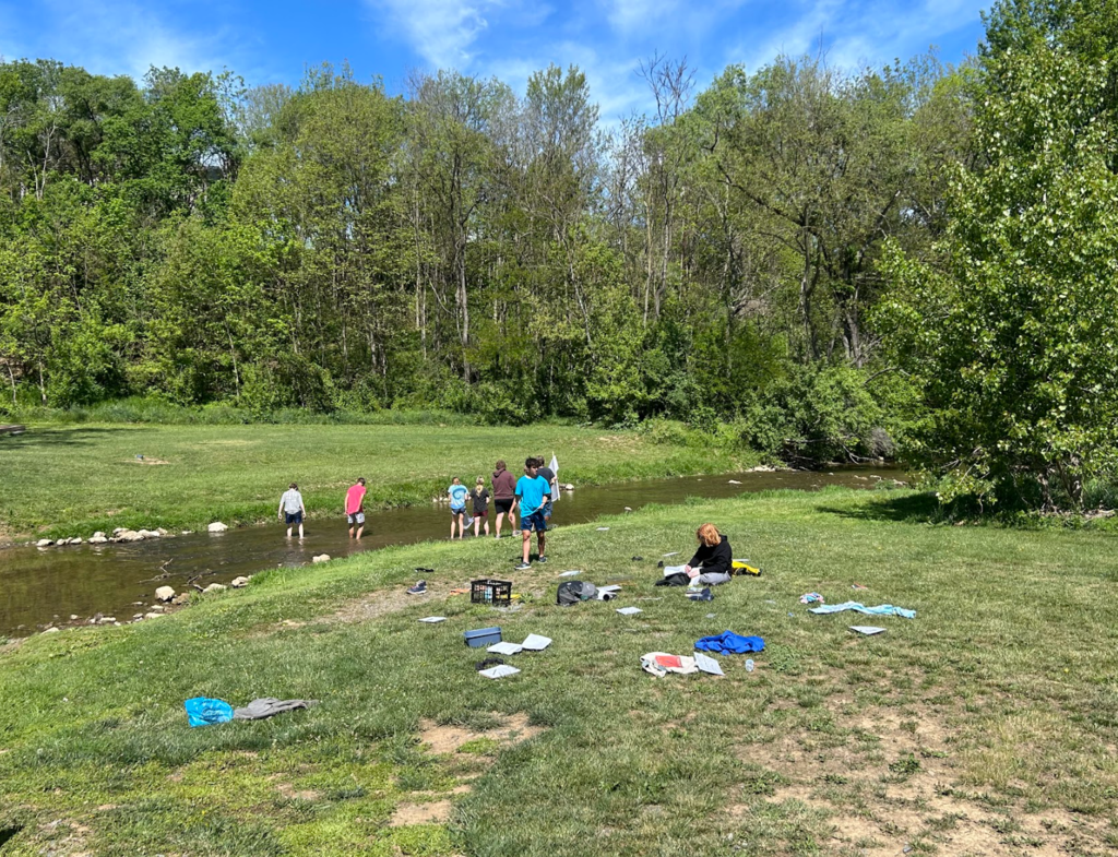 A group of people in a grassy area by a stream, some using nets to catch fish.