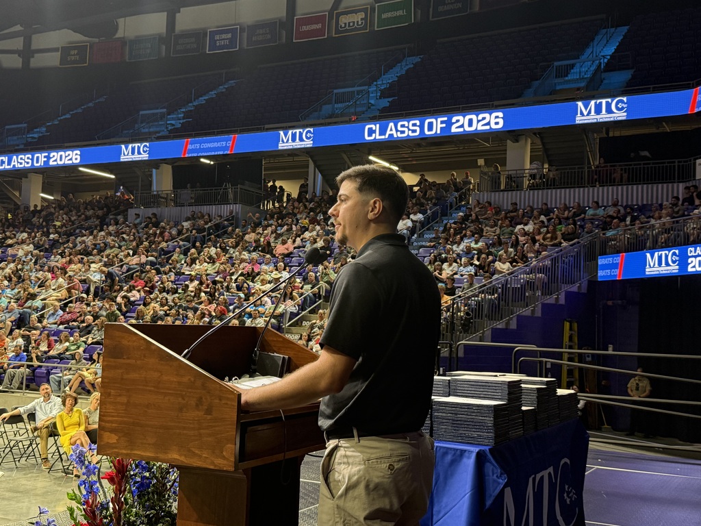 A man stands at a podium in a large auditorium, speaking into a microphone. A large crowd fills the stadium seats.