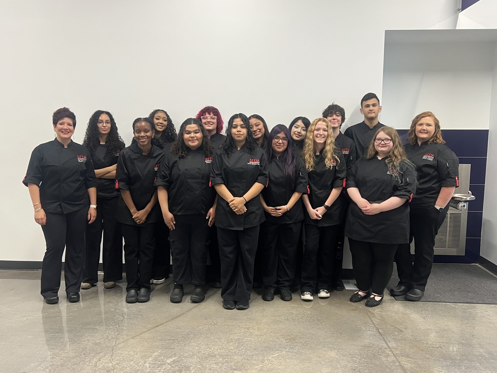 A group of people in black uniforms stands together in a room with a white wall.