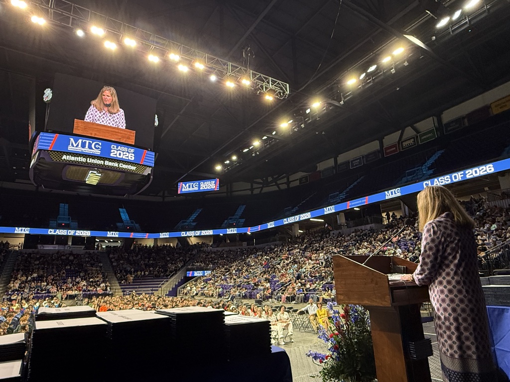Woman at podium with microphone, speaking in a large stadium, with a large video screen behind her.