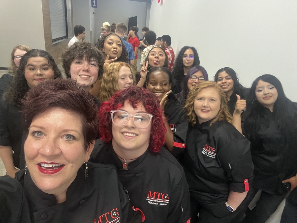 A group of women wearing black chef uniforms are posing for a selfie in a hallway.