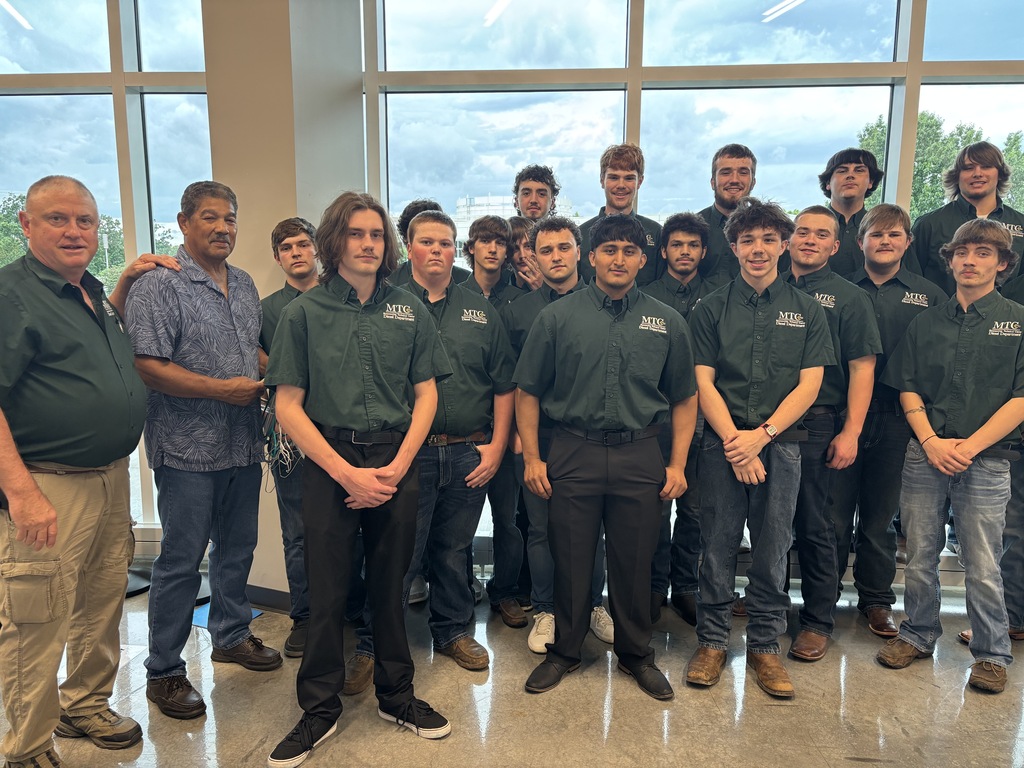 Group of young men in green shirts and pants, posing together in a large room with glass walls.