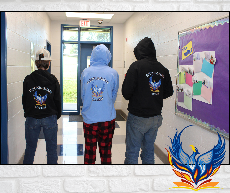 Three individuals in hoodies stand in a hallway. A bulletin board with paper pinned to it is on the wall.