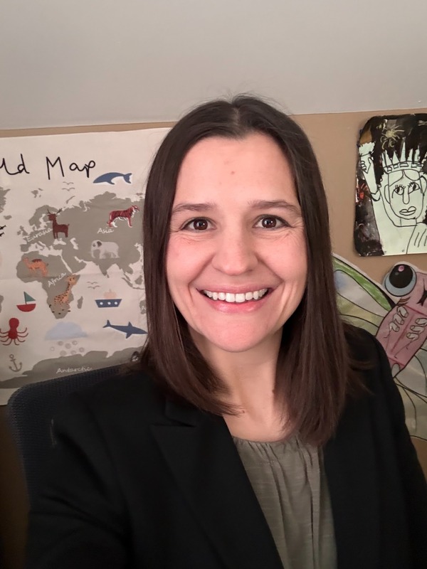 Mrs. Lindsley is pictured in front of a world map wearing a gray shirt and black jacket