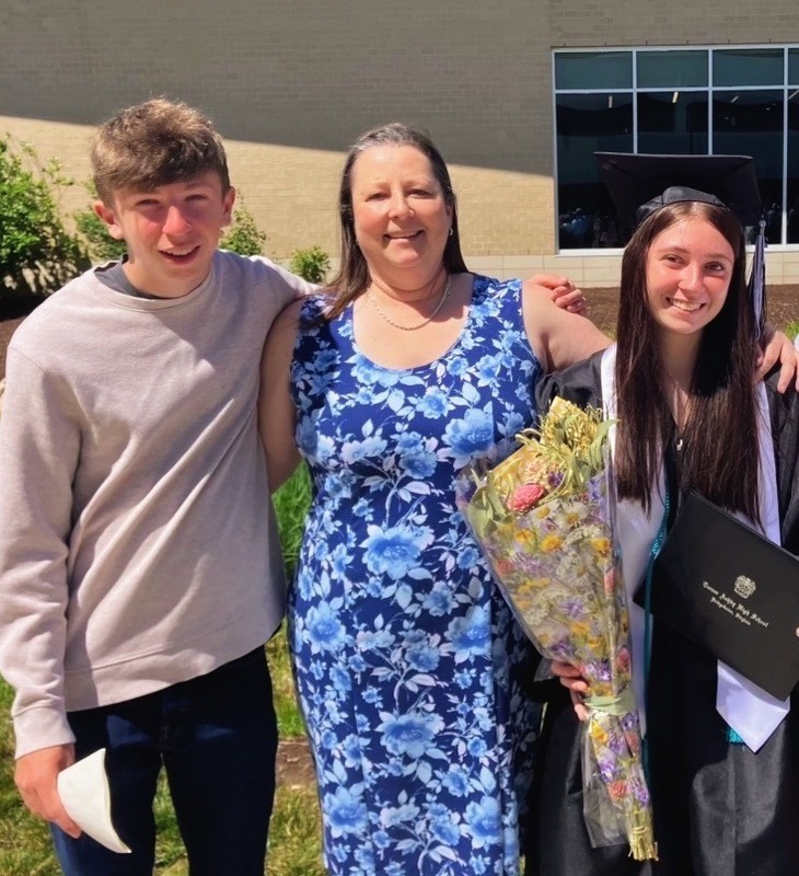 Ms. Carriker is pictured wearing a blue flowered dress. He son is to the left of her in a gray shirt and her daughter is to the right in a cap and gown holding a bouquet of flowers. 